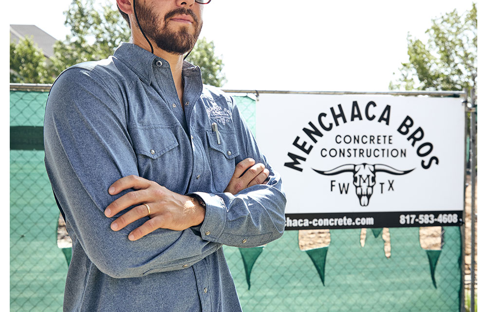 A man, Nathan Menchaca, in a blue button-up work shirt stands with arms crossed in front of a Menchaca Bros Concrete Construction sign at a job site, with fencing and trees in the background.